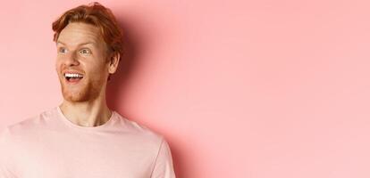 Close-up of handsome caucasian guy with red hair and beard, looking left with happy smile, checking out promo offer, standing over pink background photo