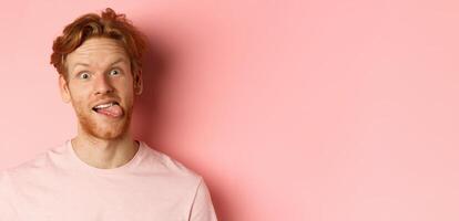 Headshot of funny redhead guy showing tongue, making silly faces at camera, standing joyful against pink background photo