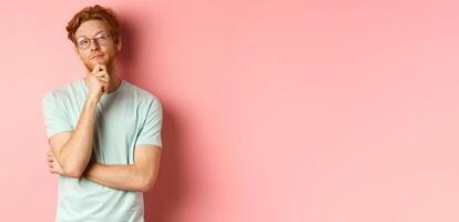 Pensive handsome redhead man in glasses looking at upper right corner, touching chin and thinking, standing over pink background photo