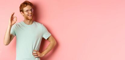 Cheerful guy with red hair and beard, wearing glasses, showing OK sign in approval and winking, smiling satisfied, standing over pink background photo