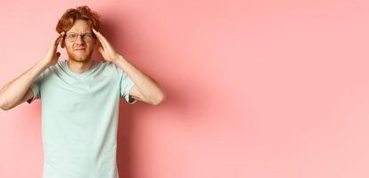 Portrait of redhead man in crooked glasses touching head and feeling dizzy or nauseous, having hangover or headache, standing over pink background photo