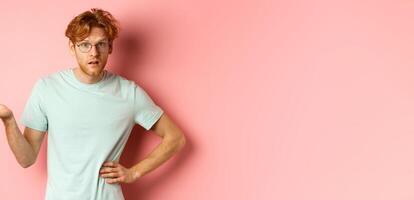 So what. Confused redhead man raising hand and shrugging, looking puzzled, dont understand something, standing over pink background photo