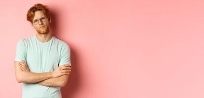 Young man with red hair and beard, wearing glasses and t-shirt, cross arms on chest, frowning while staring with skeptical and doubtful expression, standing over pink background photo