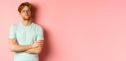 Arrogant redhead guy in glasses cross arms on chest, looking at something with skeptical face, standing over pink background photo