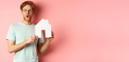 Real estate. Surprised young man with red hair and beard, wearing glasses and t-shirt, showing paper house cutout and looking impressed, saying wow, standing over pink background photo