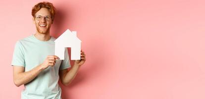 Real estate. Handsome redhead man in t-shirt and glasses, showing paper house cutout and smiling, standing over pink background photo
