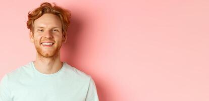 Happy young man with beard and messsy red haircut, smiling with white teeth and cheerful expression, standing over pink background photo