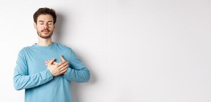 Portrait of young caucasian man with beard standing with hands on heart and closed eyes, dreaming about happy moments, standing over white background photo
