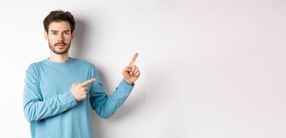 Portrait of serious caucasian man with beard pointing right at logo and looking at camera, standing in casual clothes on white background photo