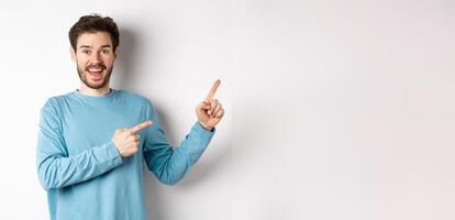 Cheerful smiling man pointing fingers right and showing banner or logo on white space for advertisements, recommending product, standing over studio background photo