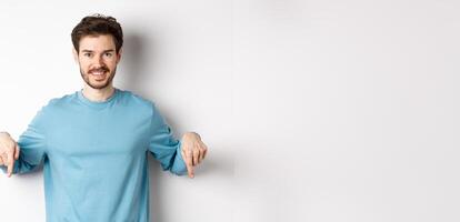 Smiling confident man pointing fingers down, showing promo banner or logo on white background, standing in casual blue sweatshirt photo