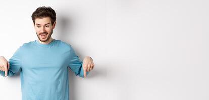 Excited and happy young man looking and pointing down at awesome deal, checking out promotion offer, standing on white background photo