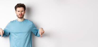 Sulking man pointing fingers down at something bad, frowning and complaining, standing over white background photo
