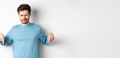 Skeptical boy pointing fingers down and frowning disappointed, looking at bad promo offer, standing on white background photo