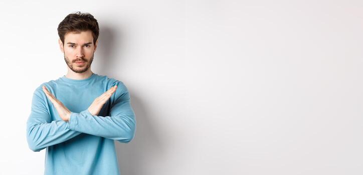joven hombre con barba mirando grave, haciendo cruzar gesto a detener algo, prohibir acción, en pie en contra blanco antecedentes foto