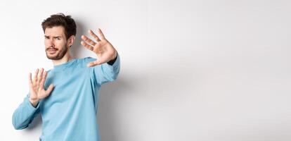 Stay away from me. Reluctant young man step back with hands stretch out in defensive gesture, protecting himself, standing over white background photo