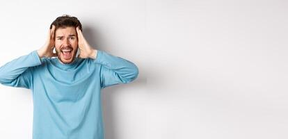 Image of young caucasian man screaming in panic, looking frustrated and shouting at camera, standing on white background photo
