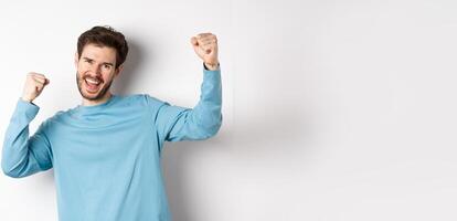 Image of happy young man triumphing, feeling like champion, winning and screaming in cheer, standing over white background photo