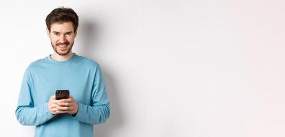 Smiling modern man using smartphone and looking pleased at camera. Guy in sweatshirt with mobile phone on white background photo