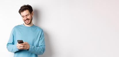 Portrait of modern caucasian man with beard, wearing casual clothes, reading smartphone screen and smiling, networking via mobile phone, standing over white background photo
