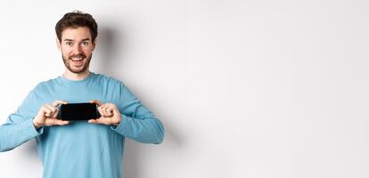 E-commerce and shopping concept. Smiling cheerful man showing empty smartphone screen in horizontal position, looking happy at camera, white background photo