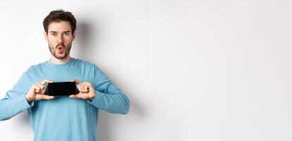 E-commerce and shopping concept. Image of impressed man saying wow and showing blank mobile phone screen in horizontal, standing over white background photo