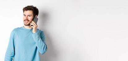 Technology concept. Young male model talking on mobile phone, calling someone on smartphone and smiling, standing over white background photo