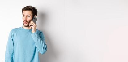 Technology concept. Intrigued young man looking left impressed, talking on mobile phone, calling someone, standing over white background photo