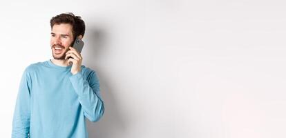 Technology concept. Joyful man enjoying phone call, talking on smartphone and smiling, standing in casual shirt over white background photo