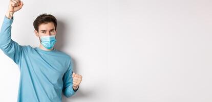 Covid-19, pandemic and social distancing concept. Happy and lucky guy in medical mask winning, raising hands up and celebrating achievement, white background photo