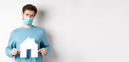 Covid and real estate concept. Sad and doubtful young man in medical mask feeling reluctant, showing paper home cutout, standing over white background photo