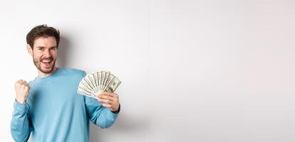 Happy man dancing with money, showing dollars and saying yes with satisfied smile, making fist pump gesture, standing over white background photo