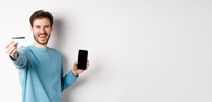 E-commerce and shopping concept. Happy man showing plastic credit card and screen of smartphone, recommending banking app, standing on white background photo