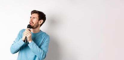 Worried and confused man reading lyrics on karaoke, looking left with unsure face, holding microphone and singing, white background photo
