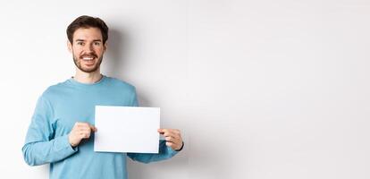 Young man with happy face showing blank piece of paper for your logo or sign, smiling at camera, standing over white background photo