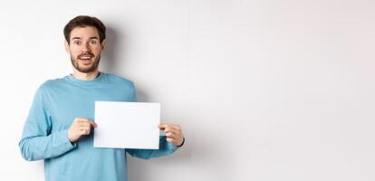 Romantic man looking with hopeful face at camera, showing sign card, blank piece of paper for banner or logo, standing over white background photo