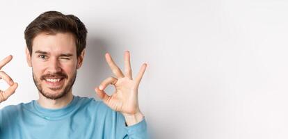 Close up of handsome bearded man winking and smiling, showing okay signs, assure all good, standing on white background photo