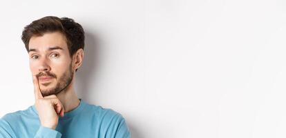 Image of thoughtful young man with beard, making choice, touching lip and looking pensive at camera, deciding something, white background photo