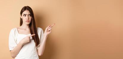 Hesitant young woman look unsure, pointing fingers right while making decision, frowning with puzzled face, standing on beige background photo