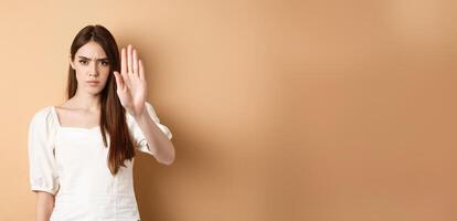 Nope stop right there. Serious and confident woman stretch out hand to forbid something, frowning and saying no, disagree and reject bad offer, standing on beige background photo