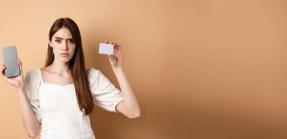 Sad and disappointed girl showing empty smartphone screen and plastic credit card, complaining and frowning, standing on beige background photo