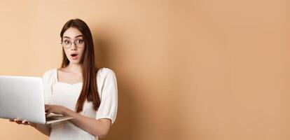 Surprised girl using laptop and gasping amazed, say wow, looking in glasses at camera, beige background photo