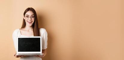 Happy woman in glasses showing laptop screen and smiling, demonstrate internet promo deal, standing on beige background photo