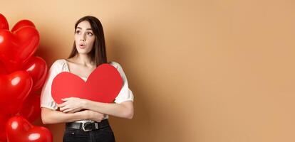 Valentines day and love concept. Intrigued tender girl hugging big red heart cutout and stand near balloons, looking aside at logo, saying wow, beige background photo