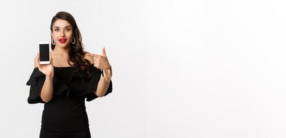 Online shopping concept. Fashionable woman in black dress pointing finger at smartphone screen, showing application, standing over white background photo