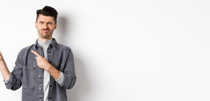 Skeptical and disappointed young man pointing fingers left, grimacing and shaking head in rejection, dislike bad product, standing on white background photo