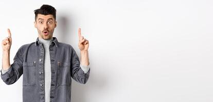 Excited man checking out promo offer, gasping amazed and pointing fingers up, making cool announcement, standing on white background photo
