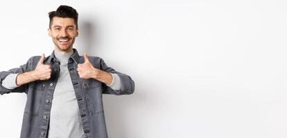 Handsome smiling man showing thumbs-up in approval, praise good work, satisfied with nice choice, recommending something cool, standing on white background photo