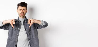 Disappointed guy showing thumbs down and frowning, looking skeptical at something bad, dislike and disapprove awful thing, standing on white background photo