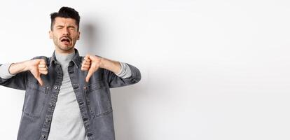 Sad man crying and showing thumbs down, whining and complainin, being upset and disappointed, standing on white background photo